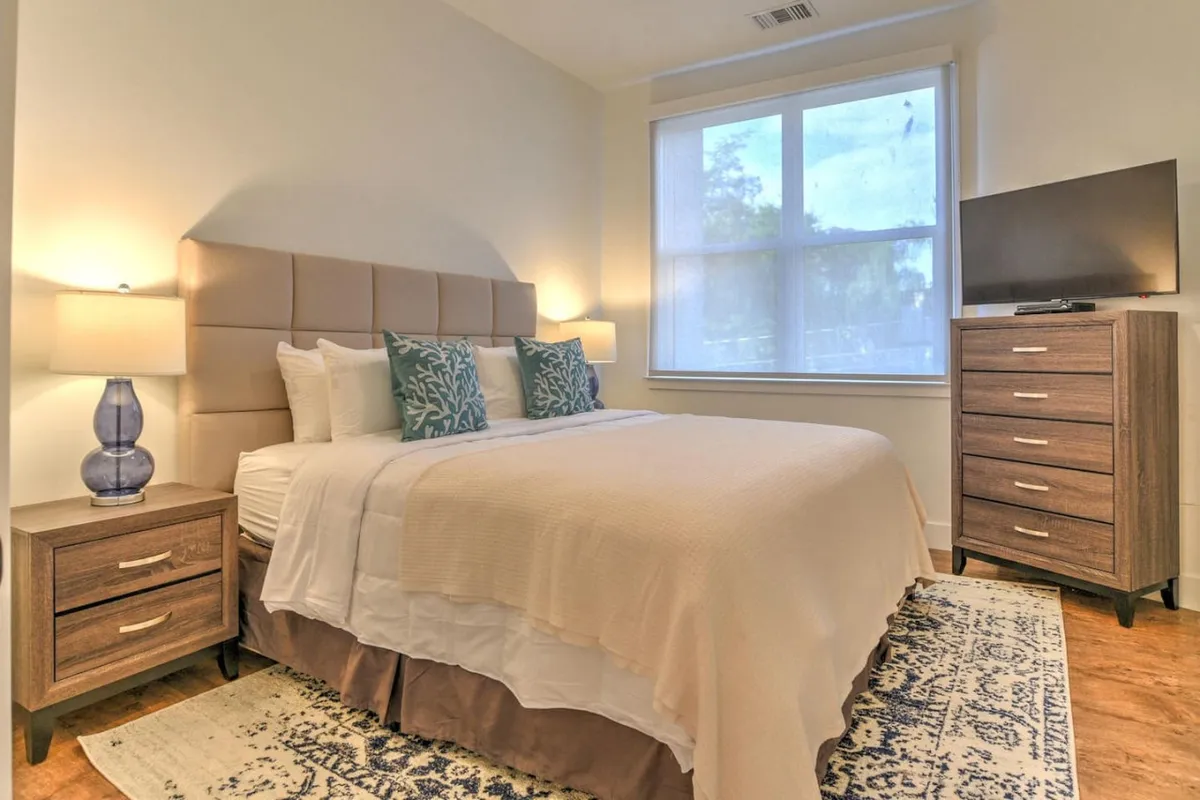 A stylish king bedroom with a large bed topped with inviting decorative pillows and a cozy throw. The room includes two nightstands, each with its own lamp, and a wall-mounted television, all set against a backdrop of tasteful, modern décor. The subtle color palette and clean lines create a tranquil, inviting atmosphere.