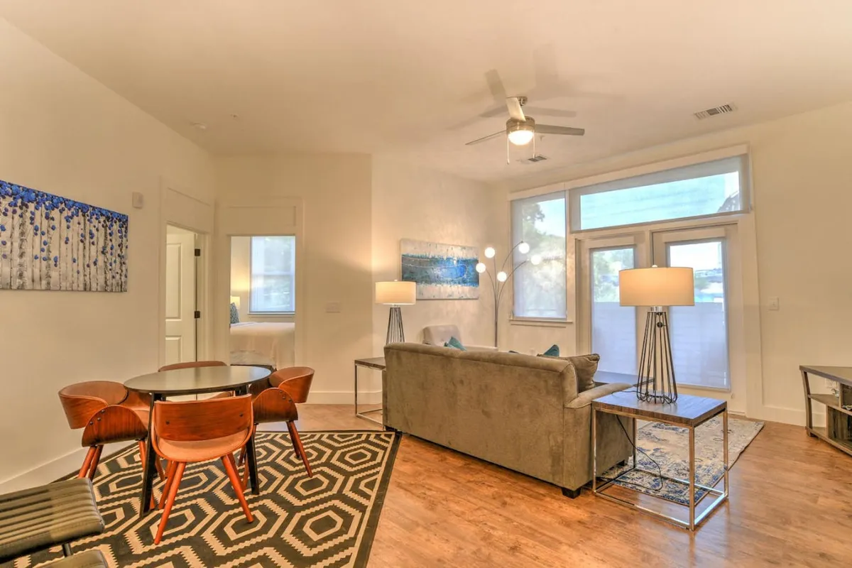 The image showcases a stylish, light-filled living and dining area featuring a round dining table perfect for gatherings, vibrant abstract artwork that adds a modern touch, a plush sofa inviting relaxation, and a cozy floor lamp ideal for reading or unwinding. The space feels open and welcoming, with hints of natural light streaming in, suggesting a comfortable spot for morning coffee or evening conversations.