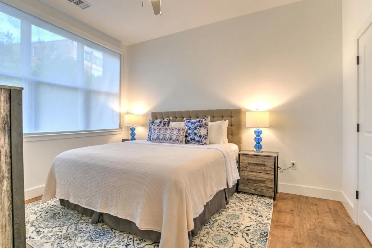 A peaceful bedroom retreat featuring a king-sized bed with layered, textured linens and decorative pillows. The space includes a nightstand with a lamp and a soft area rug underfoot. Large windows invite daylight, enhancing the room's calm and airy feel, while contemporary accents complete the look.