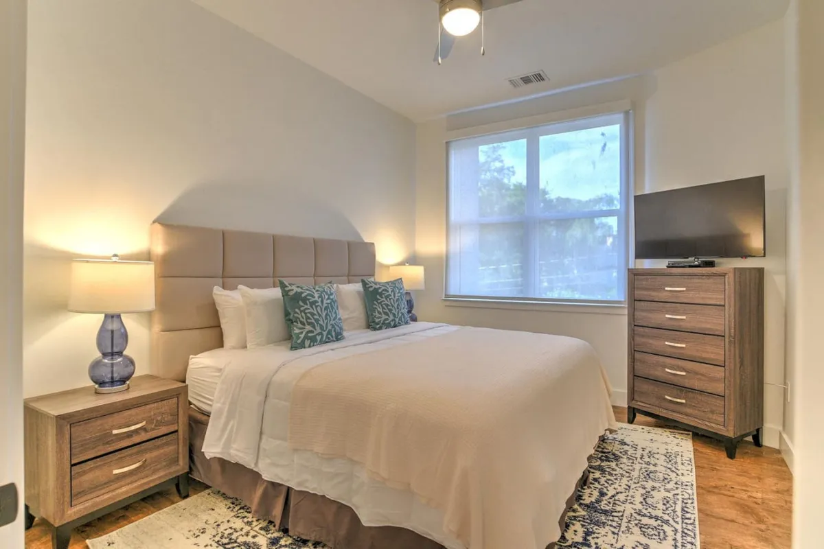 A serene, well-lit bedroom featuring a king-sized bed adorned with plush decorative pillows and crisp linens. Matching nightstands with lamps flank the bed, providing balance and convenience, while a window allows soft natural light to fill the space. A dresser with a TV sits across from the bed, combining comfort and entertainment in a contemporary setting.